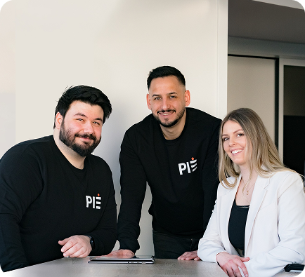 Drei PIE Marketing Teammitglieder am Tisch, zwei im schwarzen PIE Pullover, eine im weißen Blazer, Bürohintergrund